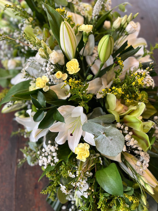 casket spray of flowers with white lilies, green leaves, and yellow accents on a wooden surface.