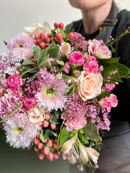 A close-up of a hand-held floral bouquet featuring pink and white roses, purple chrysanthemums, green foliage, and clusters of small pink berries. A person wearing a black top is partially visible holding the bouquet against a plain light background.