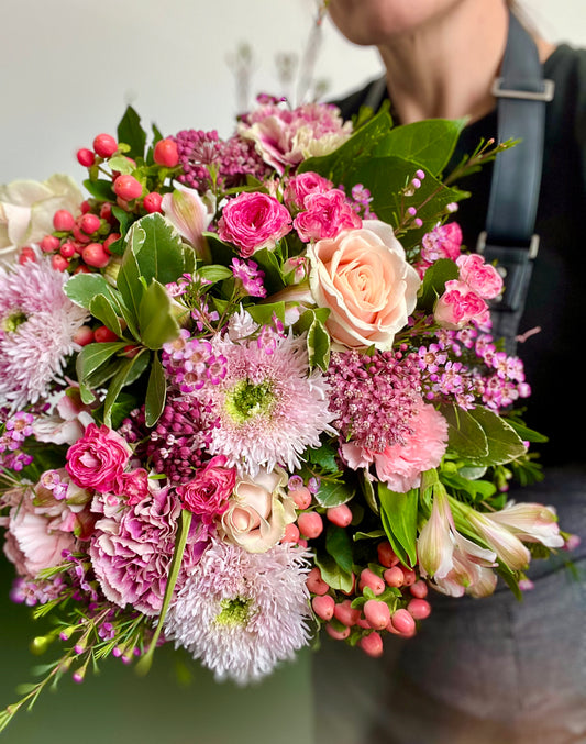 A close-up of a vibrant floral bouquet featuring pink roses, purple chrysanthemums, pink berries, and green foliage, held by a person wearing a black apron.